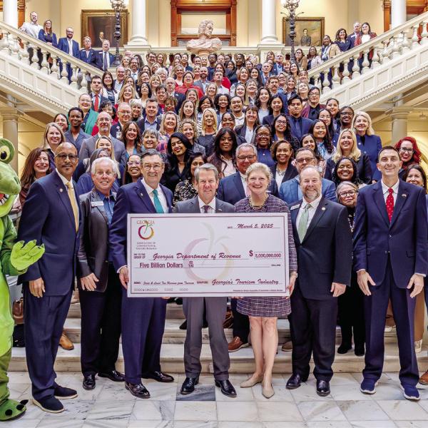 Gov. Brian Kemp (front center) accepts a symbolic $5 billion check from Amanda Dyson-Thornton, executive director of the Georgia Association of Convention &amp; Visitors Bureaus. MMCVB Executive Director Jennifer Rosa León is at top right of the main staircase before it turns right. CONTRIBUTED